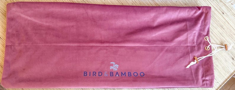 May include: A dusty rose-colored velvet bag with a drawstring closure. The bag features the text "BIRD & BAMBOO" in a dark blue font. The drawstring is white with wooden beads. The bag is rectangular in shape.