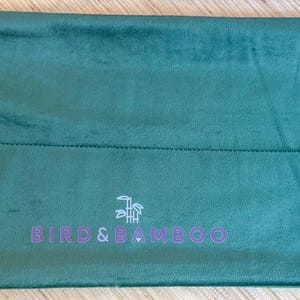 May include: A teal green, rectangular bag with a drawstring closure. The bag has a stitched seam across the middle and the text "BIRD & BAMBOO" in purple. The drawstring is white.
