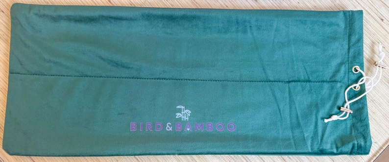 May include: A teal green, rectangular bag with a drawstring closure. The bag has a stitched seam across the middle and the text "BIRD & BAMBOO" in purple. The drawstring is white.