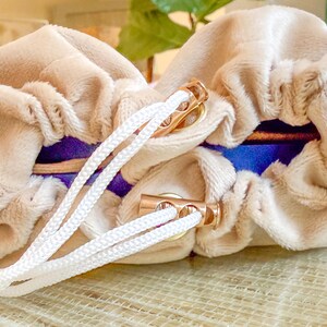 May include: Two beige velvet pouches with white drawstrings and gold-colored metal accents. The pouches have a blue and purple interior. The drawstrings are tied together. The pouches are likely for storing jewelry or small items.