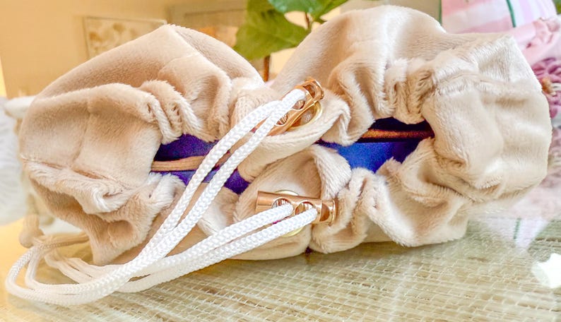 May include: Two beige velvet pouches with white drawstrings and gold-colored metal accents. The pouches have a blue and purple interior. The drawstrings are tied together. The pouches are likely for storing jewelry or small items.