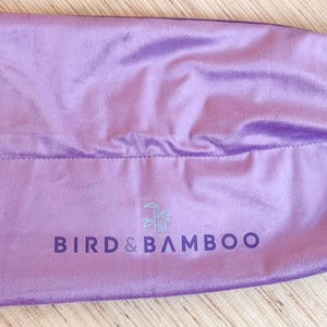 May include: A lavender satin drawstring bag with the text "BIRD & BAMBOO" printed in dark purple. The bag is closed with a white drawstring and a small gold-colored clasp.
