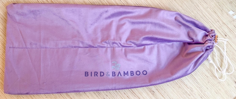 May include: A lavender satin drawstring bag with the text "BIRD & BAMBOO" printed in dark purple. The bag is closed with a white drawstring and a small gold-colored clasp.