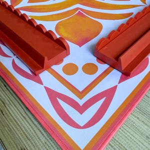 May include: An orange and white patterned play mat with two orange plastic track pieces. The mat features a geometric design with orange, pink, and white colors. The track pieces have a scalloped edge.