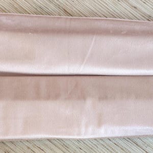 May include: A light pink satin drawstring bag, folded in half. The bag is closed with a white drawstring. The fabric has a soft, velvety appearance. The bag is lying on a light wood surface.