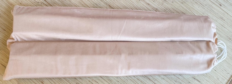 May include: A light pink satin drawstring bag, folded in half. The bag is closed with a white drawstring. The fabric has a soft, velvety appearance. The bag is lying on a light wood surface.
