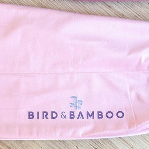 May include: A light pink drawstring bag with the text "BIRD & BAMBOO" in dark purple. The bag has a white drawstring closure and a small logo of a bird and bamboo. The bag is made of a soft, smooth material.