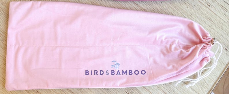 May include: A light pink drawstring bag with the text "BIRD & BAMBOO" in dark purple. The bag has a white drawstring closure and a small logo of a bird and bamboo. The bag is made of a soft, smooth material.