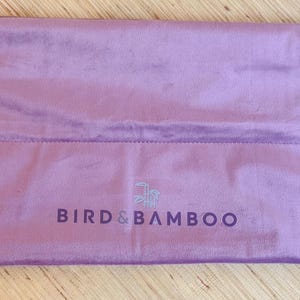 May include: A lavender-colored, rectangular drawstring bag made of a soft, velvety material. The bag features the text "BIRD & BAMBOO" in a darker shade of purple. The drawstring is white with gold-colored metal eyelets.