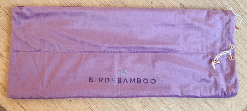 May include: A lavender-colored, rectangular drawstring bag made of a soft, velvety material. The bag features the text "BIRD & BAMBOO" in a darker shade of purple. The drawstring is white with gold-colored metal eyelets.