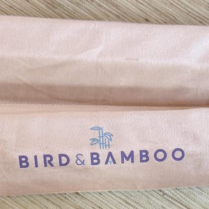 May include: Two light pink, cylindrical fabric cases with drawstrings, lying side by side. The cases have the words "BIRD & BAMBOO" printed in dark purple, with a bamboo graphic above the text. The cases are made of a soft, possibly velvet-like material.