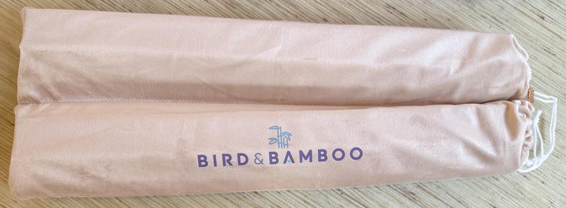May include: Two light pink, cylindrical fabric cases with drawstrings, lying side by side. The cases have the words "BIRD & BAMBOO" printed in dark purple, with a bamboo graphic above the text. The cases are made of a soft, possibly velvet-like material.