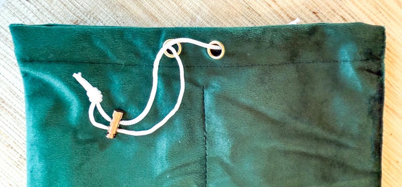 May include: A close-up of a dark green velvet drawstring bag. The bag has a white drawstring with a wooden toggle. The top of the bag has two gold-colored grommets. The bag is made of a soft, plush material.