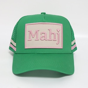 May include: Green trucker hat with a curved brim and mesh back. The front features a light pink rectangular patch with the word "Mahj" embroidered in pink. Pink and white stripes accent the sides.