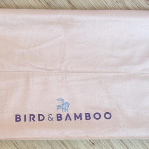 May include: A light pink Bird & Bamboo drawstring bag. The bag is rectangular with a seam across the middle and the brand name printed in dark blue. White drawstring cords with wooden accents are visible on the right side.