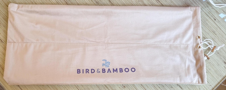May include: A light pink Bird & Bamboo drawstring bag. The bag is rectangular with a seam across the middle and the brand name printed in dark blue. White drawstring cords with wooden accents are visible on the right side.