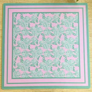 May include: A pink square placemat with a green border and a repeating pattern of white flowers on a pink background.