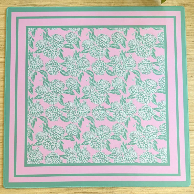 May include: A pink square placemat with a green border and a repeating pattern of white flowers on a pink background.