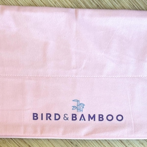 May include: A light pink Bird & Bamboo drawstring bag. The bag is made of a soft material and features the brand name in dark blue lettering. The drawstring closure has gold-colored metal accents and white cords. The bag is laying flat on a light brown surface.