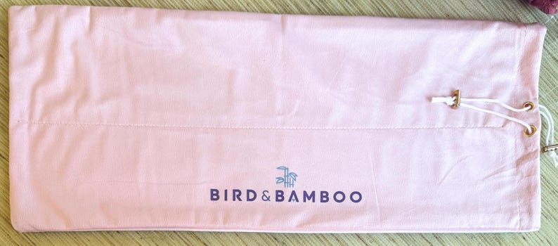 May include: A light pink Bird & Bamboo drawstring bag. The bag is made of a soft material and features the brand name in dark blue lettering. The drawstring closure has gold-colored metal accents and white cords. The bag is laying flat on a light brown surface.