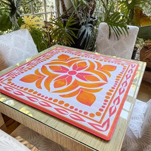 May include: A square table mat with a floral design in shades of orange and pink. The mat is placed on a square table with a glass top. The table is surrounded by white chairs with a leaf pattern.