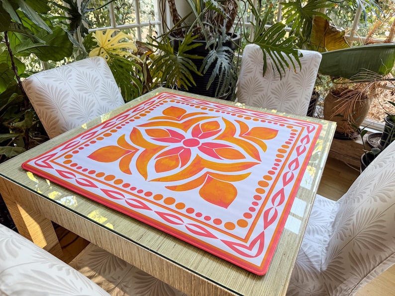 May include: A square table mat with a floral design in shades of orange and pink. The mat is placed on a square table with a glass top. The table is surrounded by white chairs with a leaf pattern.