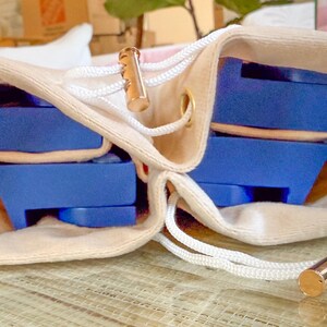 May include: A beige drawstring bag with a gold-colored metal accent and white cord. Inside the bag are several blue objects. The bag is open, revealing the contents. The objects appear to be some type of storage or organizational tool.