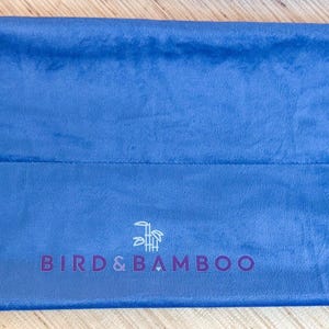 May include: A blue velvet bag with a drawstring closure. The bag has the text "BIRDS & BAMBOO" printed in purple. The bag is lying flat on a wooden surface.