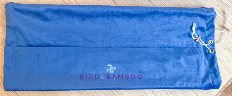 May include: A blue velvet bag with a drawstring closure. The bag has the text "BIRDS & BAMBOO" printed in purple. The bag is lying flat on a wooden surface.