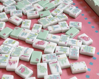 May include: A collection of white and mint green Mahjong tiles scattered on a pink surface. The tiles feature various illustrations in blue, pink, and green, including dragons, pagodas, and floral designs. Some tiles have numbers and Chinese characters.
