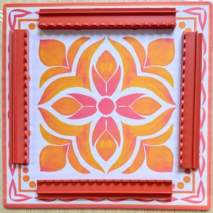 May include: A square, decorative mat with a floral design in shades of orange and pink. The mat is bordered by a red frame and includes four red, textured edge pieces. The design is centered on a white background.