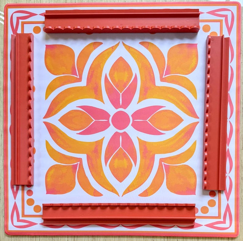 May include: A square, decorative mat with a floral design in shades of orange and pink. The mat is bordered by a red frame and includes four red, textured edge pieces. The design is centered on a white background.