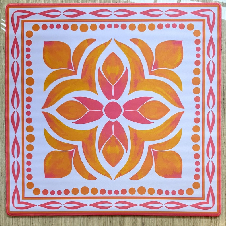May include: Square, white placemat with a floral design in shades of orange and pink. The floral pattern is centered and surrounded by a border of dots and decorative lines in orange and pink. The placemat has a square shape.