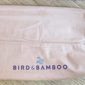 May include: A light pink, drawstring bag with the text "BIRD & BAMBOO" in dark purple. The bag is made of a soft material and has a drawstring closure with a white cord. The bag is lying flat on a light-colored surface.