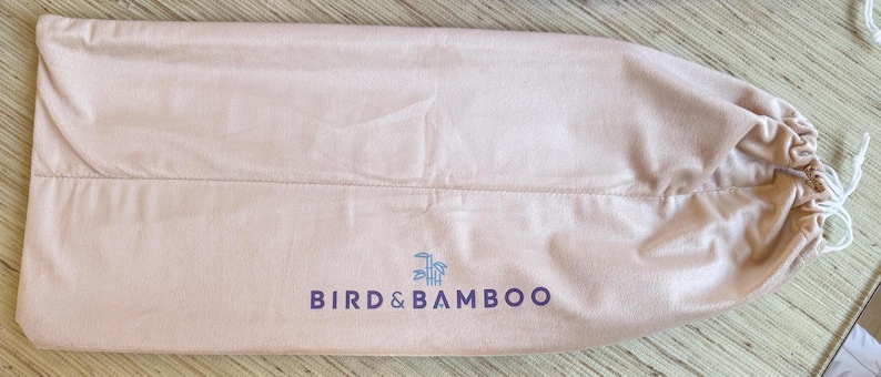 May include: A light pink, drawstring bag with the text "BIRD & BAMBOO" in dark purple. The bag is made of a soft material and has a drawstring closure with a white cord. The bag is lying flat on a light-colored surface.