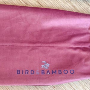 May include: A dusty rose-colored drawstring bag made of a soft, velvety material. The bag features the text "BIRD & BAMBOO" in a dark blue font, along with a small logo. The drawstring is white.
