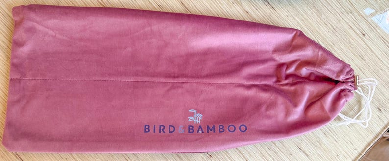 May include: A dusty rose-colored drawstring bag made of a soft, velvety material. The bag features the text "BIRD & BAMBOO" in a dark blue font, along with a small logo. The drawstring is white.