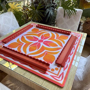 May include: A square game board with a floral design in orange and pink, framed by red edges and four matching red game pieces. The board sits on a table with a gold-colored surface, surrounded by chairs.