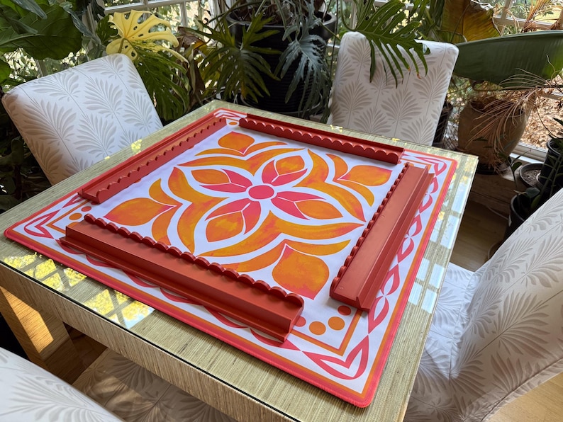 May include: A square game board with a floral design in orange and pink, framed by red edges and four matching red game pieces. The board sits on a table with a gold-colored surface, surrounded by chairs.