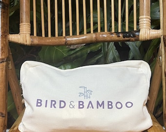 May include: A cream-colored pillow with the text "BIRD & BAMBOO" in purple, along with a bamboo graphic. The pillow is resting on a bamboo chair with a natural finish. The background features green foliage.