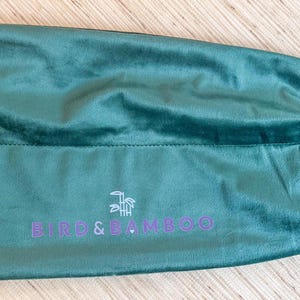 May include: A teal green velvet drawstring bag with the text "BIFU & BAMBOO" in purple. The bag is closed with a white drawstring. The bag is lying on a light brown wooden surface.