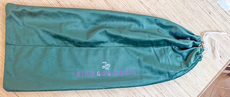 May include: A teal green velvet drawstring bag with the text "BIFU & BAMBOO" in purple. The bag is closed with a white drawstring. The bag is lying on a light brown wooden surface.