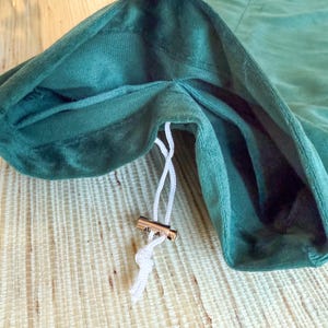 May include: Close-up of a dark green velvet drawstring bag. The bag's opening is visible, revealing a lighter green interior. A white drawstring with a gold-colored metal clasp is partially pulled, suggesting the bag is closing. The bag is on a light wood surface.