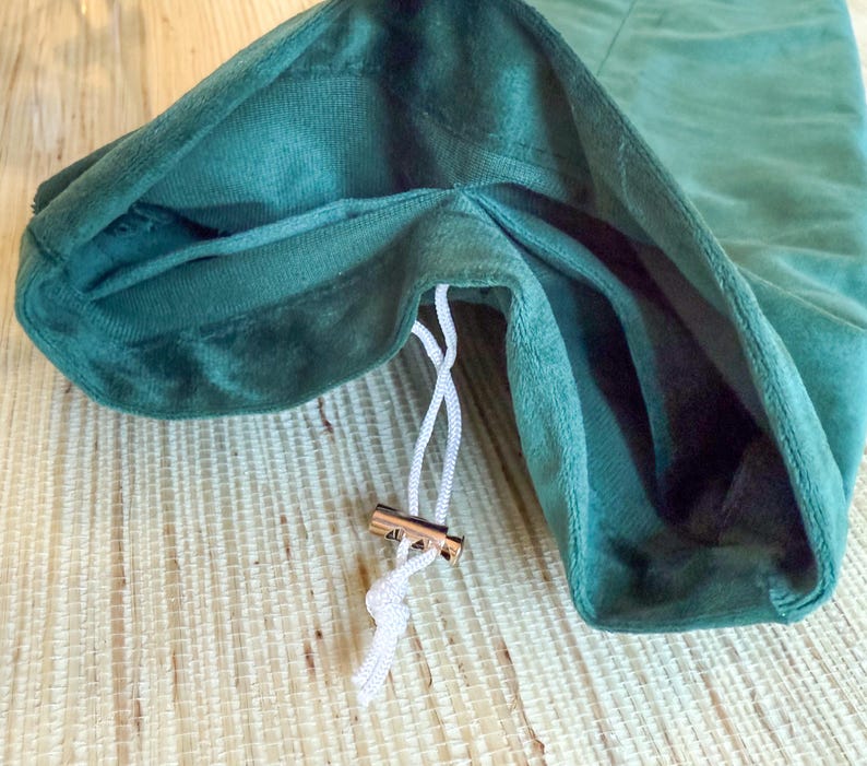 May include: Close-up of a dark green velvet drawstring bag. The bag's opening is visible, revealing a lighter green interior. A white drawstring with a gold-colored metal clasp is partially pulled, suggesting the bag is closing. The bag is on a light wood surface.