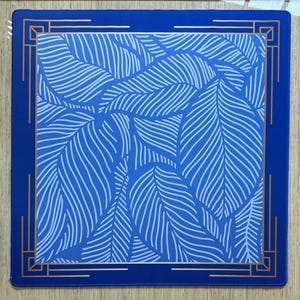 May include: A square blue placemat with a white leaf pattern. The placemat has a gold border with geometric designs and a solid blue border. The leaf pattern is a repeating design.