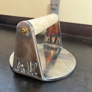 May include: A silver metal burger press with a wooden handle. The press has the letters "A1E" cut out of the metal.