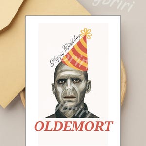 Op de afbeelding: Een witte kaart met een cartoon afbeelding van Lord Voldemort die een rood en gele feestmuts draagt. De tekst "Happy Birthday" is in zwarte cursieve letters boven de afbeelding geschreven. De tekst "OLDEMORT" is in rood onder de afbeelding geschreven.