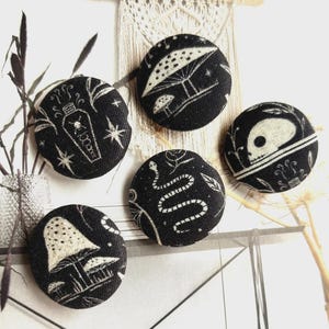 May include: A collection of five black fabric-covered buttons with white illustrations. Designs include a coffin, mushrooms, a snake, a skull, and stars. The buttons are round and have a textured appearance, suitable for sewing or crafting projects.