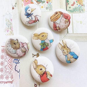 Peut inclure: Ensemble de six boutons ronds recouverts de tissu, illustrant des personnages d'une histoire pour enfants. Les boutons ont des fonds blancs et représentent des animaux en vêtements colorés, dont un canard, une grenouille et plusieurs lapins.