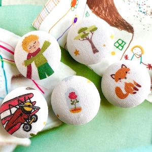 Op de afbeelding: Een verzameling van vijf ronde stoffen knopen met verschillende illustraties. De ontwerpen omvatten een jongen, een boom, een vos, een rood dubbeldekker en een roos in een glazen stolp. De knopen zijn wit met kleurrijke afbeeldingen.